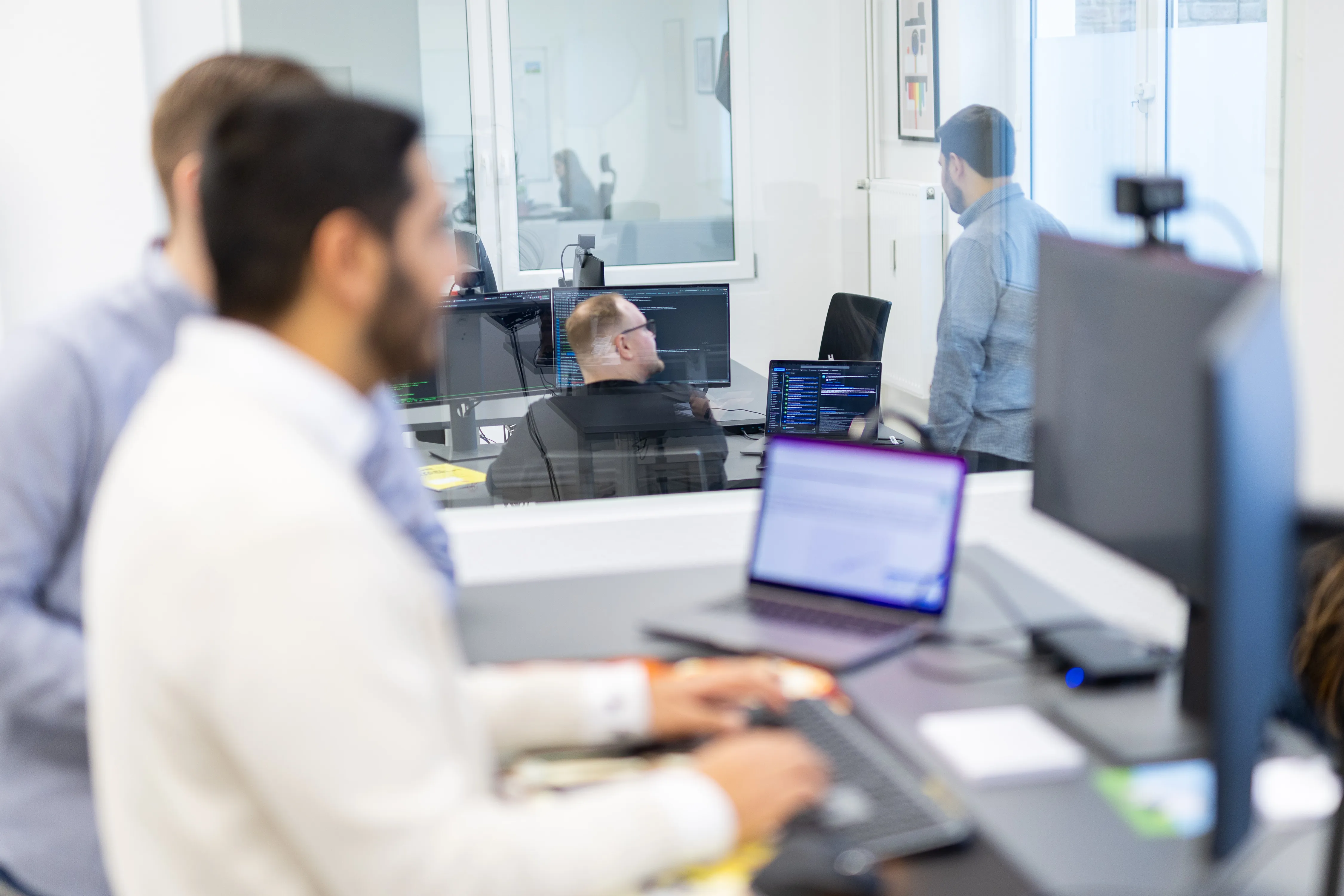 Vier Personen in einem modernen Büro mit Glaswänden. Zwei Personen arbeiten am Computer, die anderen beiden führen ein Gespräch an einem Schreibtisch.